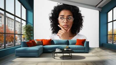 Education, people and life goals. Headshot of attractive mixed race schoolgirl wearing stylish round eyeglasses having doubtful and indecisive look, holding her chin as she has to choose college Wall mural