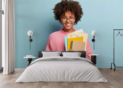 Education and ethnicity concept. Optimistic glad dark skinned young woman with pleased expression, holds textbooks nad papers, rejoices learning new information, isolated over blue background Wall mural