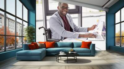 Elegant happy dark skinned male invalid looks over some paperwork, sit at work space next to big window, keyboards information on laptop computer. People, business, occupation, ethnicity concept Wall mural