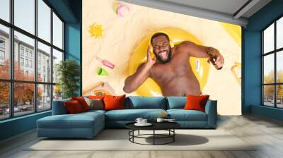 Emotional bearded guy with dark skin thick beard listens music with wireless headphones drinks beer lies on swimring at sandy beach exclaims from joy enjoys summer time. Lazy day at seaside. Wall mural