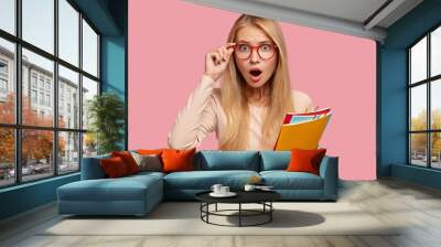 Emotional bugged eyed young blonde girl cant believe in sudden news, opens mouth widely, expresses wonder, wears transparent glasses, holds textbooks, stands over pink background. Omg concept Wall mural