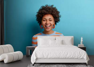 Emotions and happiness concept. Amused carefree curly haired ethnic woman has fun as attends funny show, enjoys hilarious comedy, dressed in casual clothes, poses indoor over blue studio wall Wall mural