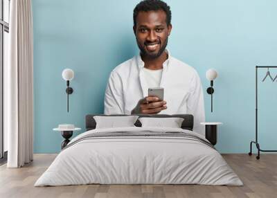 Ethnic man recieves email about discounts, uses application for publication, holds modern cell phone, dressed in white shirt, connected to wireless internet, waits for sms message, smiles positively Wall mural