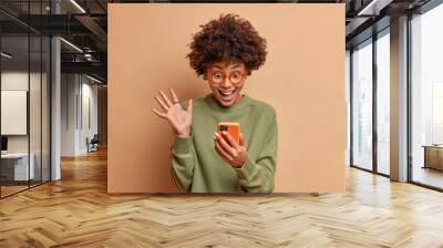 Excited cheerful dark skinned curly young woman raises palm and stares overjoyed at smartphone display being social media blogger reads amusing article or watches funny video communicates online Wall mural