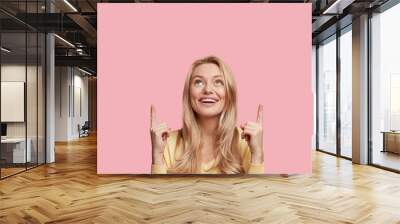 excited female with pleasant smile, long light hair, dressed in yellow sweater indicates with fore f Wall mural