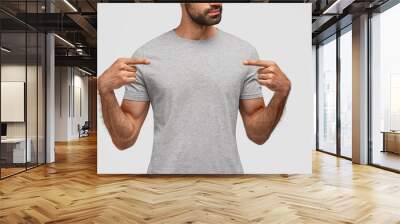 Fashion, clothing and design concept. Serious bearded young male points with two fore fingers at blank t shirt, shows free space for your advertisement or logo, isolated over white background Wall mural