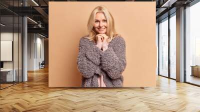 Fashionable blonde forty years old woman keeps hands under chin and smiles gently wears warm winter coat has minimal makeup poses against brown studio background. People and fashion concept. Wall mural