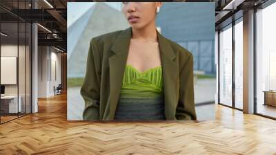 Fashionable woman with bun hairstyle dressed in stylish green clothes keeps hands in pocket looks away pensively poses at urban place during daytime. People street style and lifestyle concept Wall mural