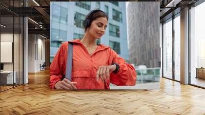 Female runner looks at smartwatch while going for run checks time listens music via headphones dressed in jacket strolls outdoor against modern tall buildings tracks wearable device wanders in city Wall mural