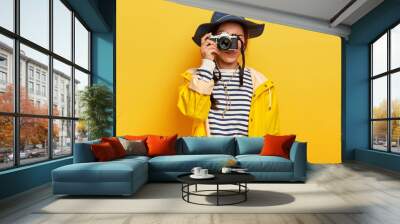 Female traveler makes memorable photos during trip, holds retro camera, takes images of beautiful landscape or place, dressed in striped jumper, raincoat and hat, isolated over yellow background Wall mural