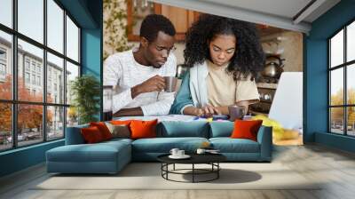 Finances and family budget concept. Indoor shot of serious African girl using calculator while paying bills, sitting at kitchen table with papers and notebook pc, doing paperwork together with her man Wall mural