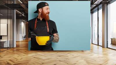 Focused unshaven man looks away, whisks something in yellow bowl, dressed in black clothes, rubber protective gloves, bakes or cooks dish, isolated over blue wall with free space. Baking concept Wall mural