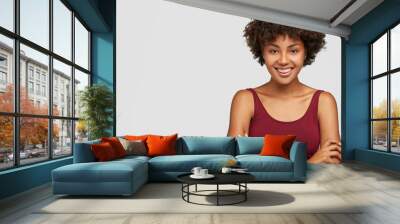 Friendly looking self assured dark skinned young girl keeps arms folded, has toothy smile, dressed in casual clothes, models against white background with free space for your promotional content Wall mural