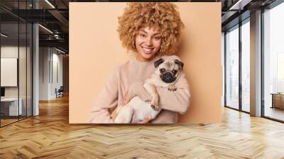 Friendship concept. Smiling beautiful young woman carries her pug dog spends free time with favorite pet wears casual jumper isolated over brown background. Domestic animal with its caring owner Wall mural