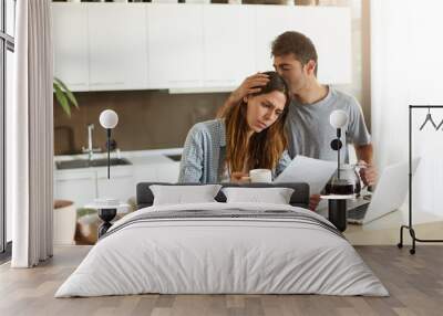 Frustrated woman studying sheet of paper in kitchen, drinking coffee and trying to find way to pay off all family debts; her husband kissing her on head, saying: Everything is going to be alright Wall mural