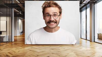 Funny attractive bearded young male with comic smile, has awkward look, pretends to be happy, looks gladfully through round spectacles, laughs at joke, isolated over white concrete background. Wall mural