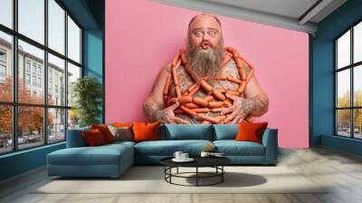 Funny fatso male keeps lips rounded, poses with naked body wrapped by sausages, has health problems connected with health and unhealthy nutrition, likes meat, stands indoor. Gluttony concept Wall mural