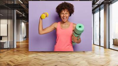 Funny happy dark skinned woman raises hand with dumbbell, shows biceps, holds rolled fitness mat, smiles broadly, wears pink casual vest, isolated over purple background. Fitness training at home Wall mural