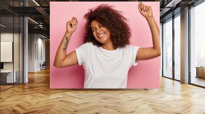 Funny joyous woman raises arms and dances carefree, feels pleasure and amused, laughs happily, eyes closed from satisfaction, moves along with music, has tattoo dressed in casual wear isolated on pink Wall mural