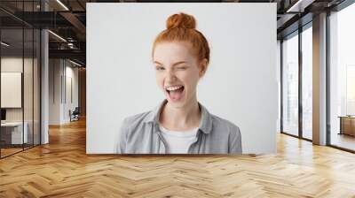 Funny red-haired female teenager with bun wearing casual shirt having joyful expression closing one of her eyes with pleasure and opening widely mouth having broad smile isolated over white background Wall mural