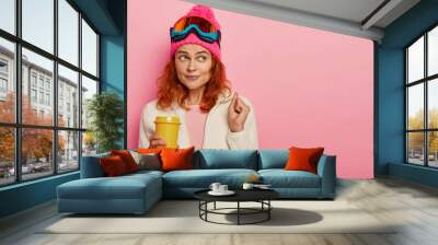 Ginger girl skier enjoys winter resort, has coffee break after reaching top of mountain, cheers dream come true, wears snowboarding glasses, warm clothing, has holidays during favourite season Wall mural