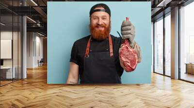 Glad cheerful butcher suggests to taste beef or pork, works on meat factory, cuts on small pieces for selling, wears special uniform, isolated over blue background. Food industry and processing Wall mural