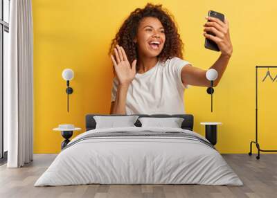 Glad dark skinned happy woman with Afro hairstyle, holds modern mobile phone in front of face, waves palm in camera, makes video call, dressed in casual wear, isolated over yellow studio wall. Wall mural