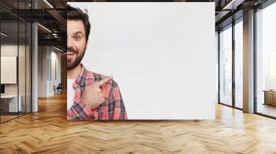 Glad smiling joyful bearded male model with trendy hairstyle, dressed in casual checkered shirt, stands against white concrete background, indicates at blank copy space for your advertising content Wall mural