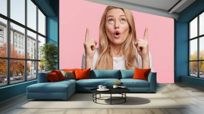 Good looking blonde female with pure healthy skin looks in amazement as indicates at something upwards, isolated over pink background. Pretty young woman sees amazing thing up, gestures indoor Wall mural