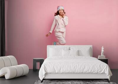 Good morning concept. Happy teenage girl in comfortable domestic clothes, jumps as awakes in good mood, has weekend or day off, models over pink background. Pretty woman in nightclothes, eyemask Wall mural