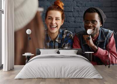 Group of three happy students having fun indoors: beautiful girl with orange hair bun sitting at cafe table close to her African boyfriend, both laughing cheerful and talking to unrecognizable female Wall mural