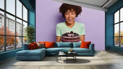 Half legth shot of female sweettooth with Afro hair, holds delicious sweet cake on plate, suggests guests taste dessert baked by herself, smiles positively, models over purple wall. Eating concept Wall mural