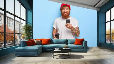 Half length shot of cheerful bearded redhead man wears stylish hat, white casual t shirt, holds mobile phone, takeout coffee, being in good mood, types messages, isolated over blue background Wall mural