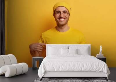 Half length shot of cheerful man points at mockup yellow t shirt, shows space for your design, has glad expression, advertises new outfit, poses against bright background. Look at this t shirt Wall mural