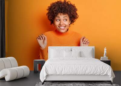 Half length shot of positive Afro American woman bites lips keeps hands raised looks curiously away smiles intrigued notices something interesting wears casual jumper isoated over orange wall Wall mural