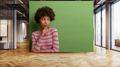 Half length shot of thoughtful dark skinned model keeps hand under chin, stands pensive against green background, thinks about future plans, wears striped jumper, focused aside, empty space. Wall mural