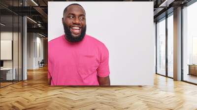 Handsome bearded man with dark skin dressed in casual pink t shirt laughs carefree shows optimism poses against grey studio wall blank copy space for your advertisement. Happy emotions concept Wall mural