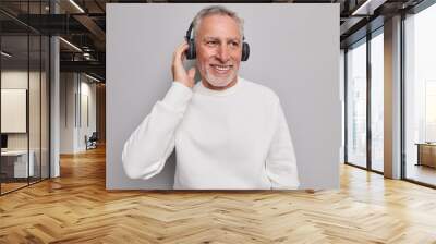 Handsome smiling mature bearded man listens audio track in wireless headphones smiles gladfully enjoys good sound quality wears neat white jumper isolated over grey background. Musical accessory. Wall mural