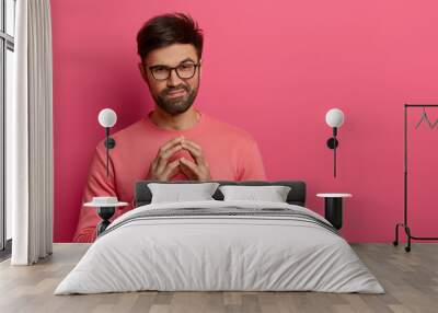 Handsome unshaven man has tricky evil plan, steepls fingers and looks mysteriously at camera, thinks over good idea, wears casual sweater and spectacles, poses over rosy background, copy space Wall mural