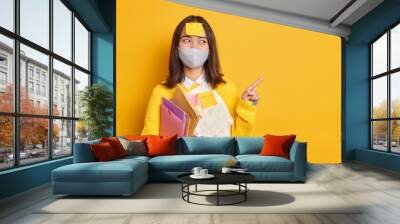 Happy Asian female manager busy doing paper work wears protective disposable mask during coronavirus pandemic indicates index finger on copy space demonstrates something over yellow background Wall mural