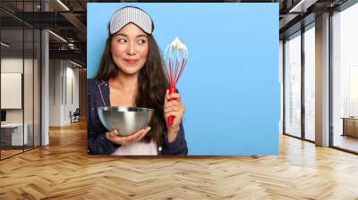 Happy busy Asian housewife whisks cream in bowl, wears sleep mask and pyjamas, cooks early in morning after aweking, poses against blue background. Woman makes homemade cake. Cooking concept Wall mural