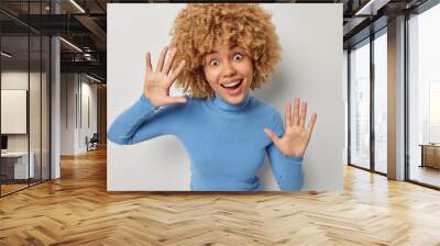 Happy cheerful curly haired European woman has surprised expression smiles broadly keeps palms raised entertains someone dressed in casual blue turtleneck poses against grey studio background Wall mural