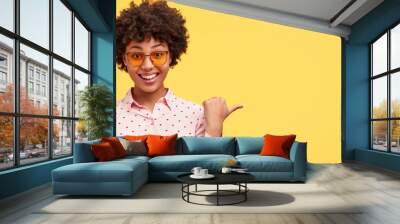 Happy delightful African American female hipster with broad smile, wears sunglasses, shows something aside with thumb, stands against yellow background. Charming dark skinned woman advertises Wall mural