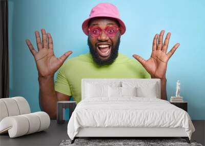 Happy emotional dark skinned bearded guy raises palms feels very glad exclaims loudly reacts on amazing news wears stylish pink sunglasses casual t shirt and panama isolated over blue background Wall mural