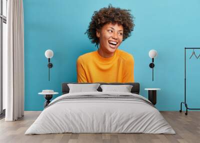 Happy emotions concept. Positive dark skinned beautiful young woman laughs poisitively looks aside with carefree face expression wears casual orange sweater isolated over blue studio background. Wall mural