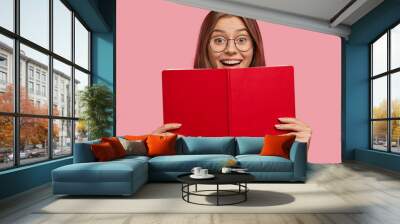 Happy European female student in spectacles, has positive expression, holds red book, rejoices successfully passed exam at university, isolated over pink background. People, learning, reading Wall mural