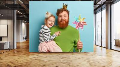 Happy ginger family of father and daughter wear party accessories, hold striped toy windmill, celebrate Fathers day or birthday, isolated over blue background. Fatherhood and celebration concept Wall mural