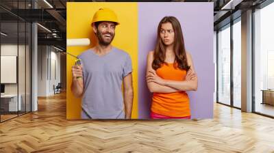 Happy hard working male in hardhat, holds paint roller, displeased wife stands crossed hands and looks at him, do house repairing at home, dressed in casual wear, isolated over two colored background Wall mural