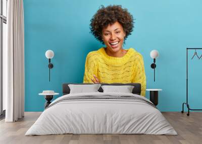 Happy joyful dark skinned woman laughs happily keeps arms folded and expresses positive emotions grins from happiness dressed in casual jumper isolated on blue background has fun or hears funny joke Wall mural