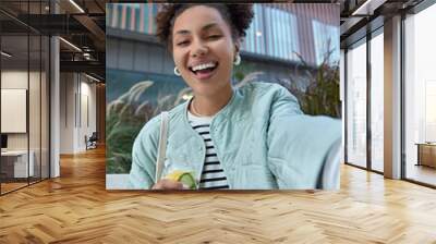 Happy optimistic girl with two hair buns dressed in jacket enjoys free time and walking in city holds bottle of detox drink makes selfie poses against modern building has fun during daytime. Wall mural